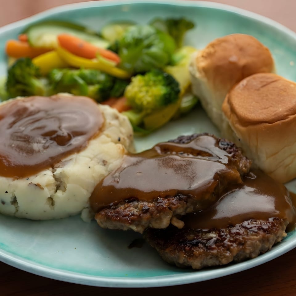 A plate of Salisbury steak with gravy, mashed potatoes, mixed vegetables, and dinner rolls.