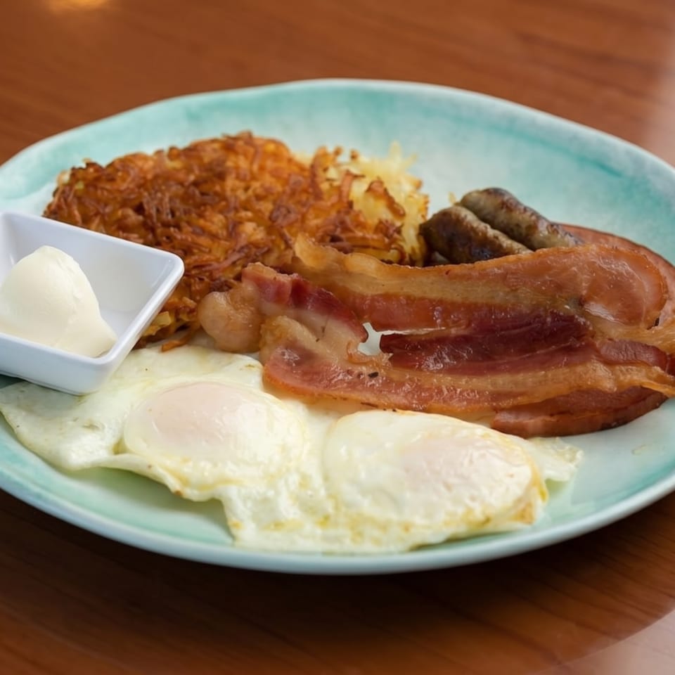 A breakfast plate with hash browns, bacon, sausage, two fried eggs, and a side of butter.