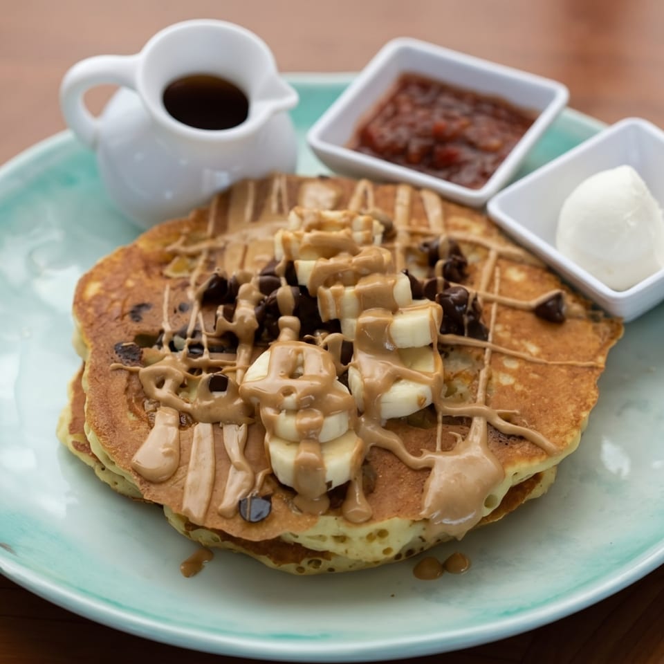 Pancakes topped with bananas, chocolate chips, peanut butter drizzle, with syrup and jam on a blue plate.